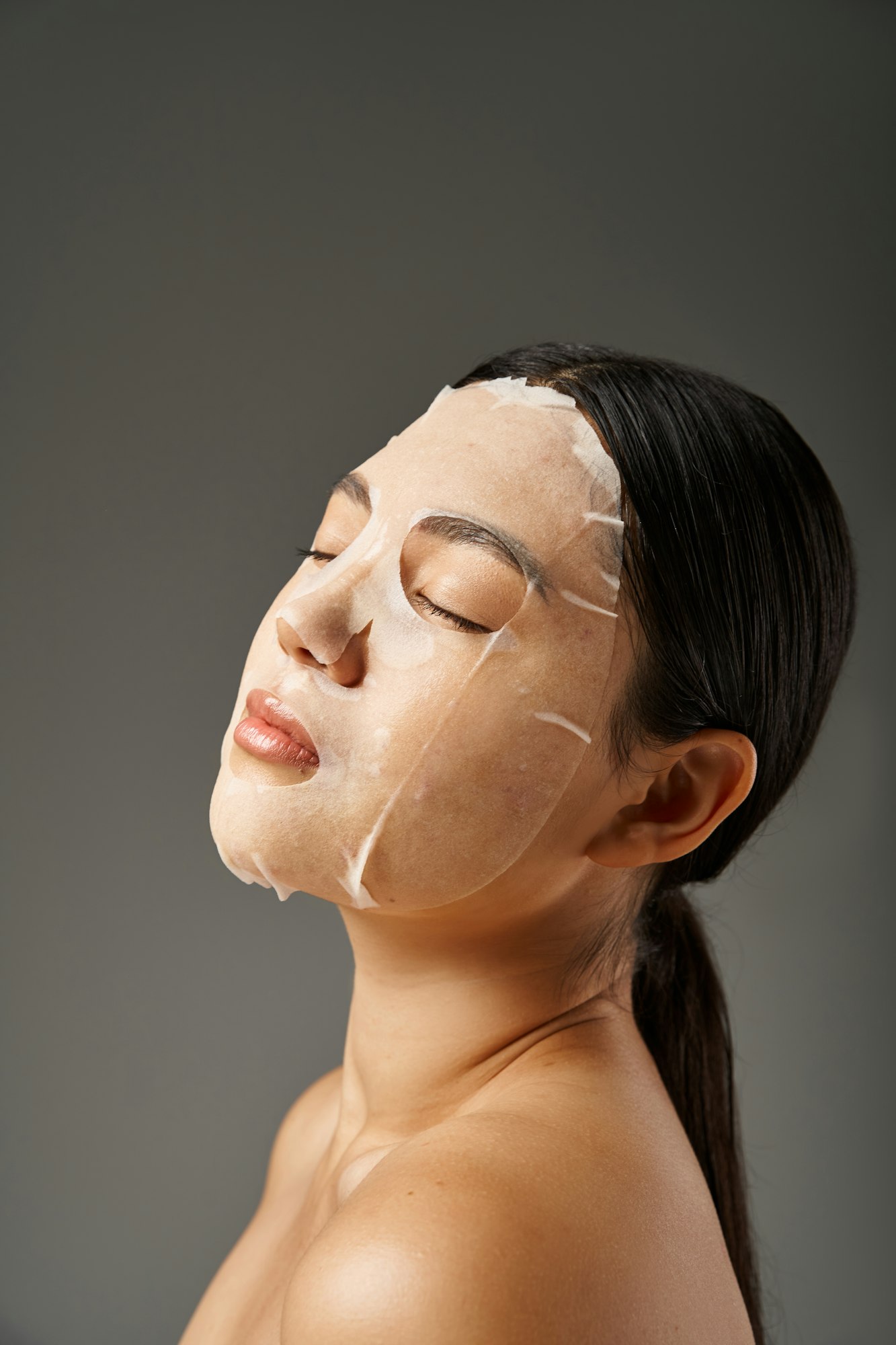 charming young asian woman with facial mask for skin treatment and closed eyes on grey background
